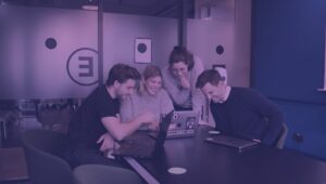 4 pessoas reunidas em uma mesa olhando fiaxamente para um notebook no centro, enquanto se olham e riem. As quatro pessoas são brancas, dois deles são homens e as doutras duas são mulheres