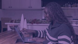 Na imagem, você confere uma mulher negra usando o notebook., A moça está de lado e sorri para a tela do computador.