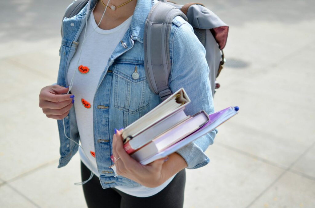 Na imagem, você vê o busto de uma mulher segurando diversos livros e usando uma mochila]!